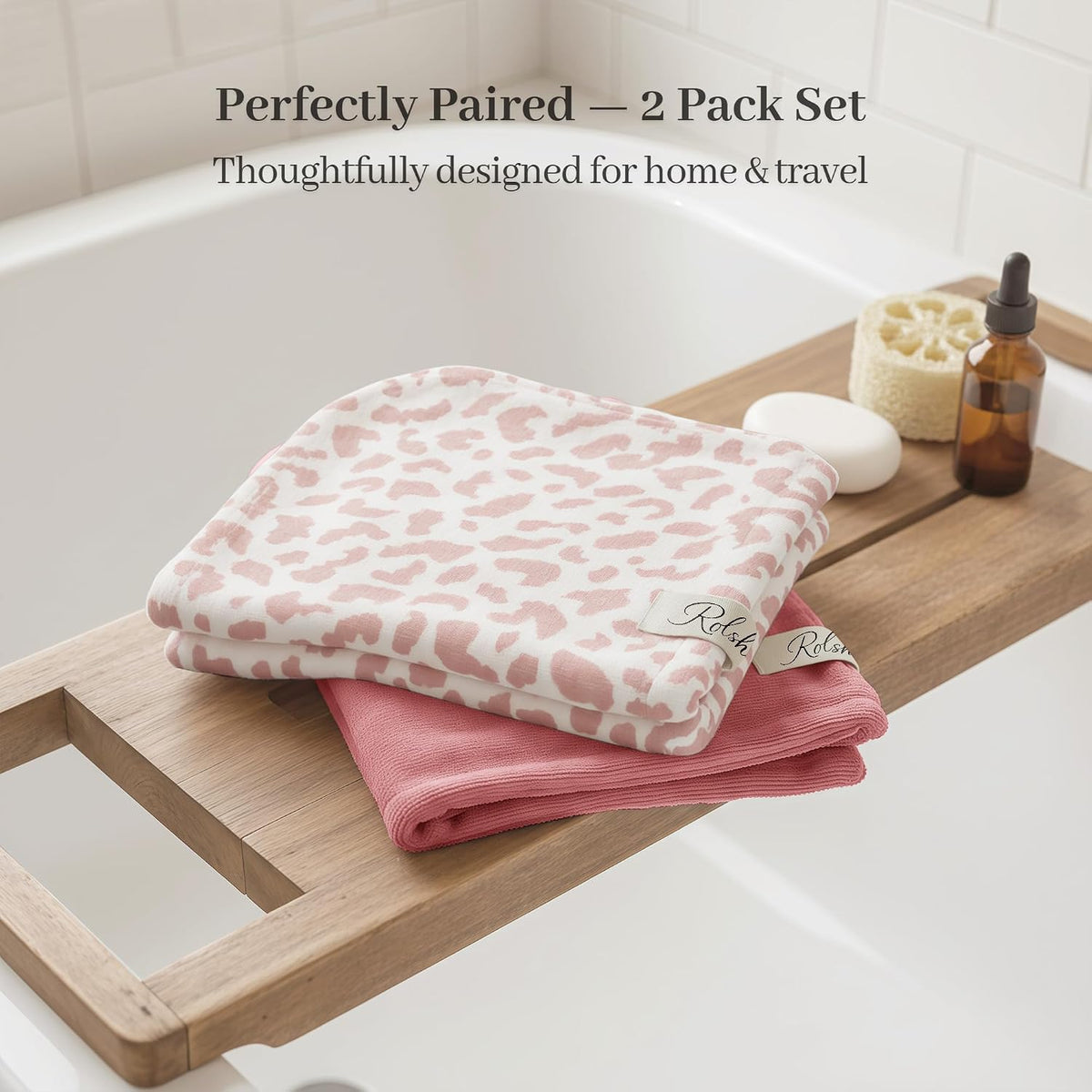 Two folded cotton hair towels on a wooden bath tray. The top towel is white with a pink leopard print pattern; the bottom is solid dusty rose. Both have small branded labels. Text reads 'Perfectly Paired - 2 Pack Set, Thoughtfully designed for home & travel.'