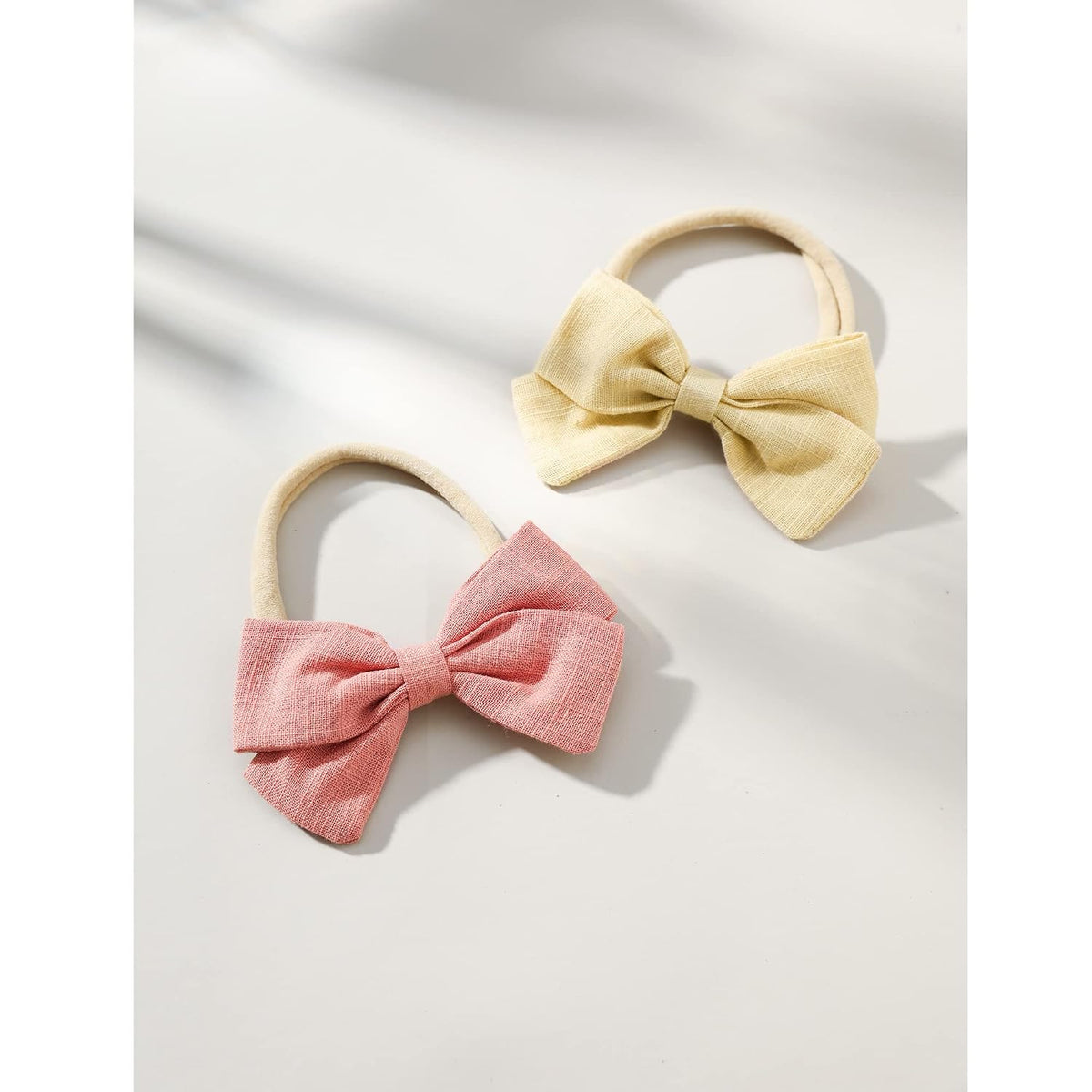 Two fabric bow hair ties on stretchy nylon bands, one dusty pink and one pale yellow, with a visible linen-like woven texture. Both bows feature a gathered center knot and wide wings, shown on a white surface.