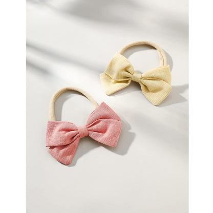 Two fabric bow hair ties on stretchy nylon bands, one dusty pink and one pale yellow, with a visible linen-like woven texture. Both bows feature a gathered center knot and wide wings, shown on a white surface.