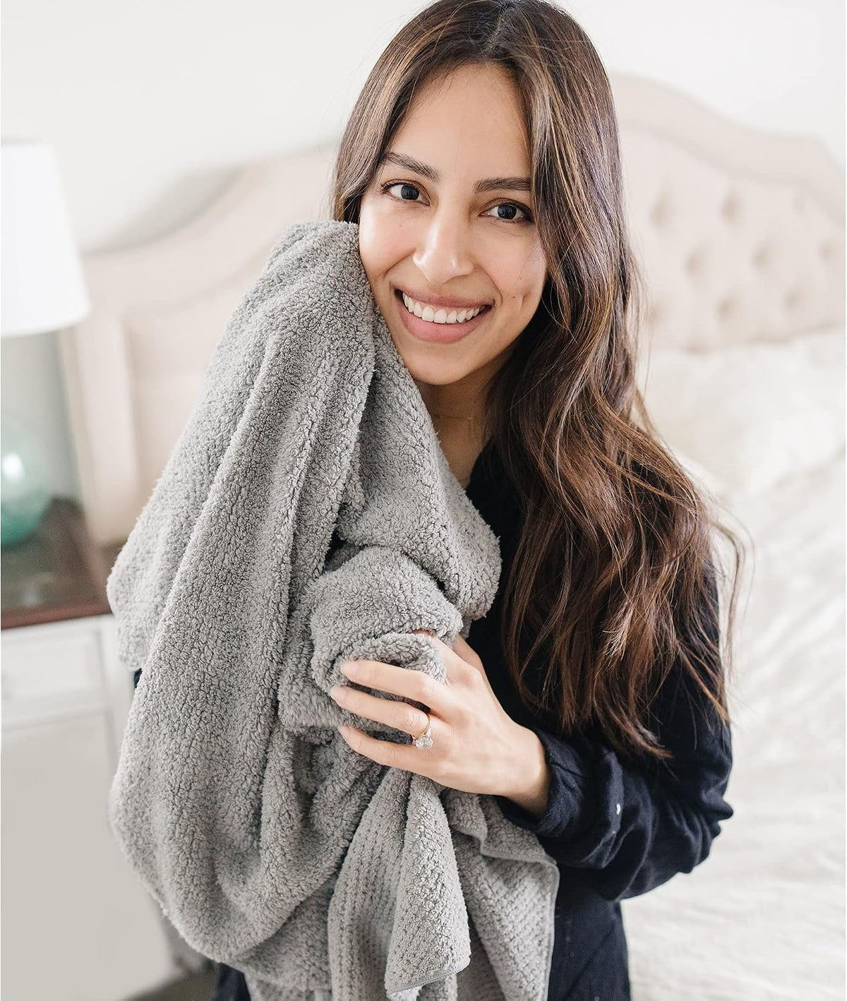 Woman with long dark hair smiling and hugging a large gray textured microfiber towel against her chest, wearing a dark top. Bedroom setting with tufted headboard visible in background.