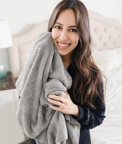 Woman with long dark hair smiling and hugging a large gray textured microfiber towel against her chest, wearing a dark top. Bedroom setting with tufted headboard visible in background.