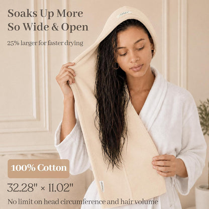 Woman in white robe wrapping a wide cream-colored cotton hair towel around her long wet dark hair. Text states 'Soaks Up More, So Wide & Open,' '25% larger for faster drying,' '100% Cotton,' 32.28" x 11.02", and 'No limit on head circumference and hair volume.'