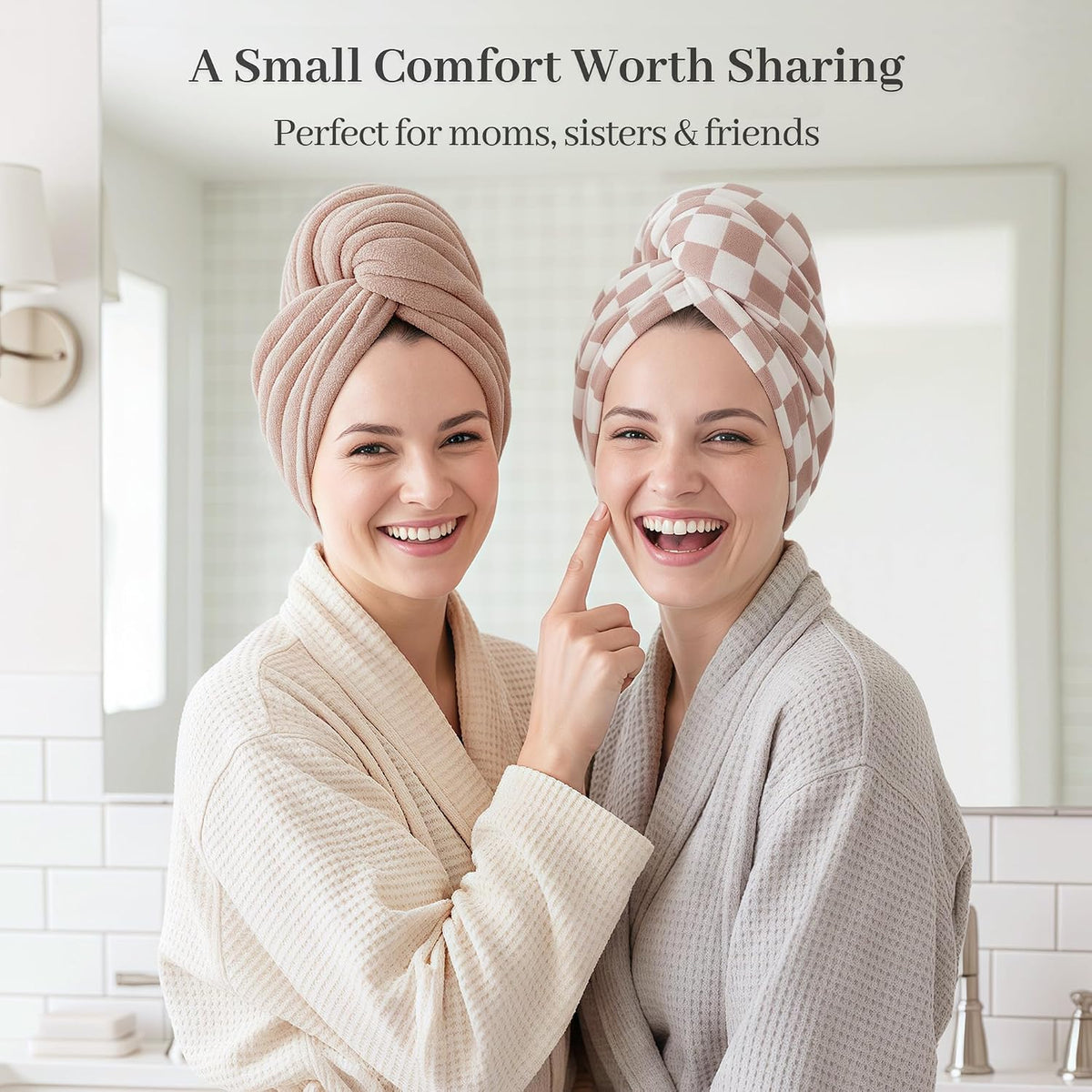 Two smiling women in waffle-weave robes wearing hair towel wraps in a bathroom setting. One wears a solid mauve wrap, the other a pink-and-white checkered wrap. Text reads: 'A Small Comfort Worth Sharing - Perfect for moms, sisters & friends'