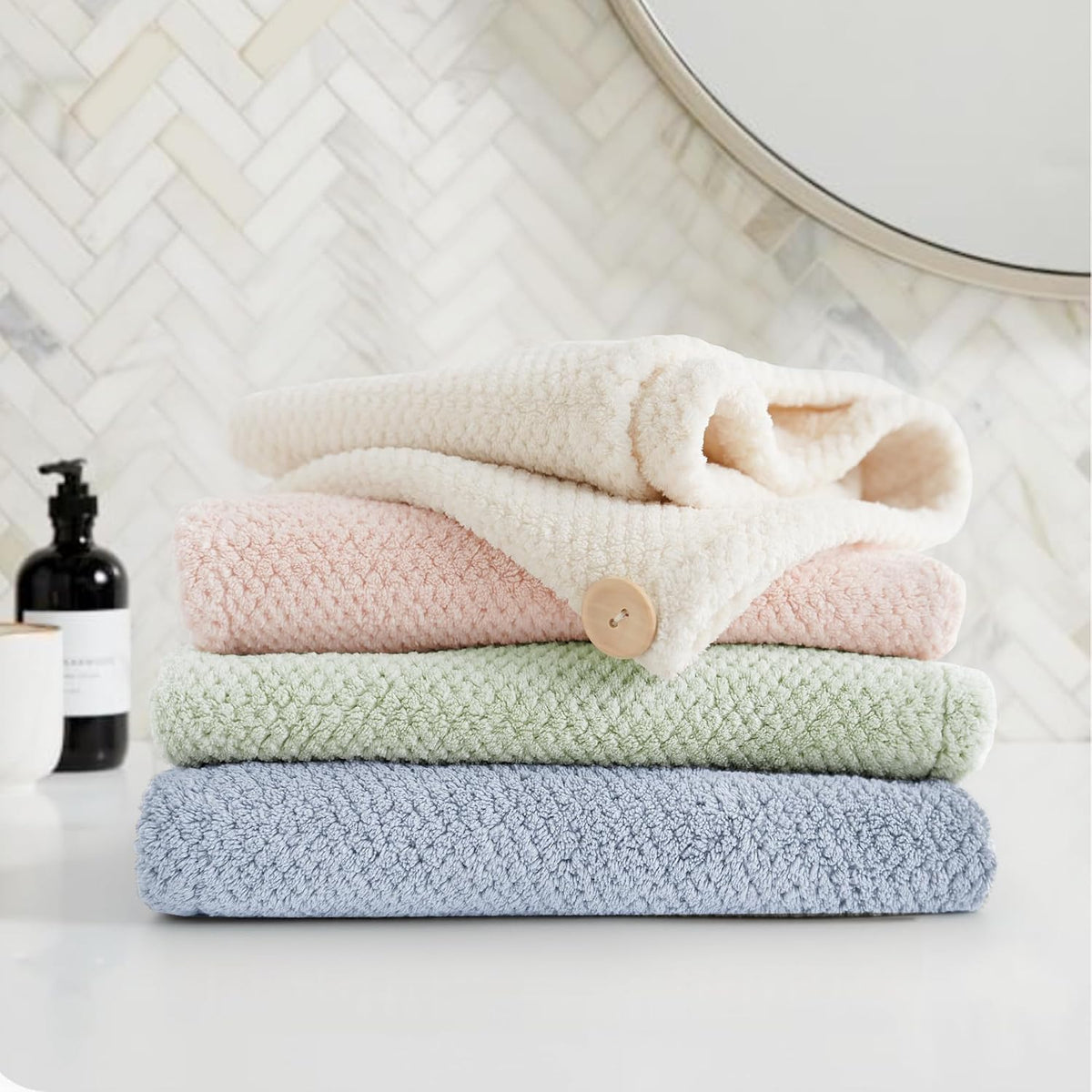 Stack of four textured microfiber hair towel wraps in blue, sage green, pink, and cream, folded and piled on a white bathroom counter. The pink towel has a visible tan button closure. Herringbone tile and a round mirror visible in background.