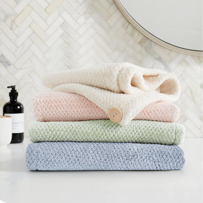 Stack of four textured microfiber hair towel wraps in blue, sage green, pink, and cream, folded and piled on a white bathroom counter. The pink towel has a visible tan button closure. Herringbone tile and a round mirror visible in background.