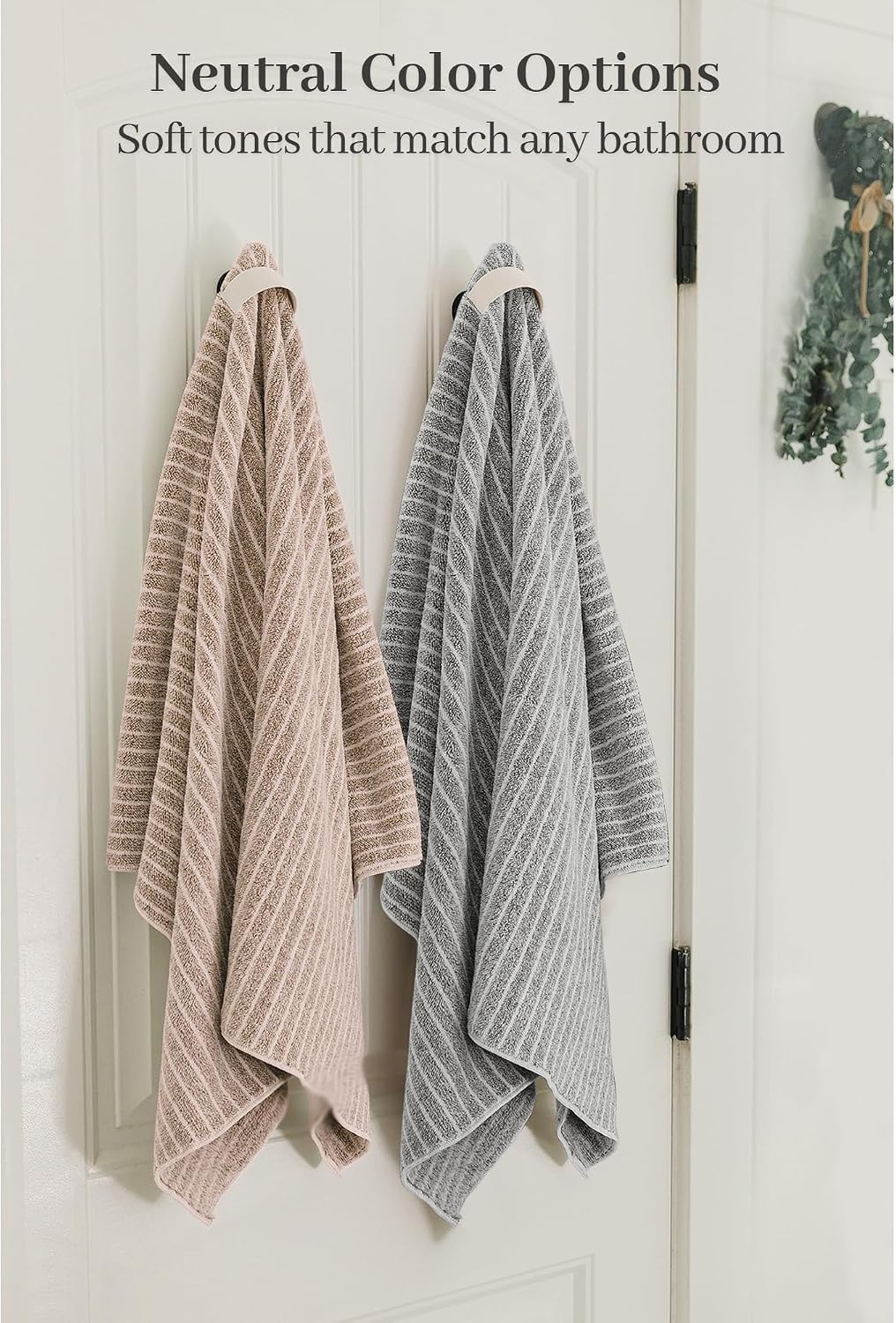 Two textured bath towels hanging on hooks on a white door -- one blush pink and one gray -- both featuring a diagonal stripe pattern. Text reads 'Neutral Color Options: Soft tones that match any bathroom.'