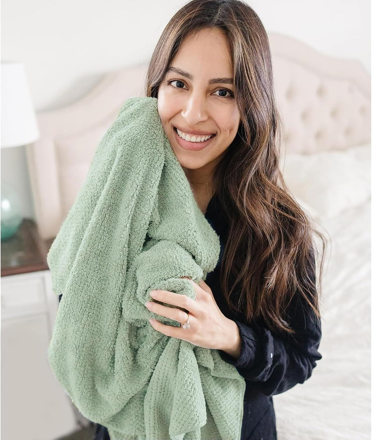 Woman in a dark top hugging a large sage green textured towel against her cheek while smiling, shown in a bedroom setting. The soft, plush towel has a waffle-like texture and drapes over her arms.