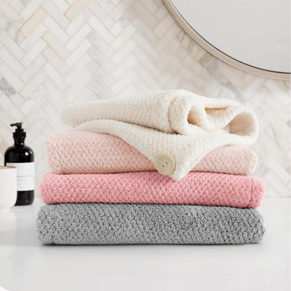 Stack of four textured microfiber hair towel wraps in gray, pink, light pink, and cream, folded and stacked on a white surface. The cream towel on top is loosely wrapped and features a small button closure. Bathroom setting with herringbone tile backdrop.