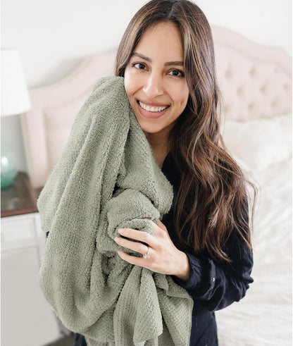 Smiling woman with long dark hair hugging a large sage green textured towel against her chest, wearing a dark top. Bedroom setting with tufted headboard visible in background.
