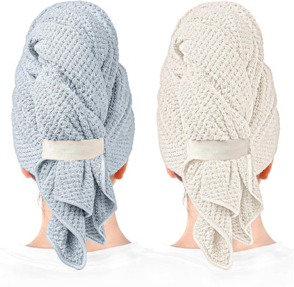 Two hair towel wraps shown worn on the backs of heads: one in muted blue and one in light beige/cream. Both feature a waffle-knit texture, a fabric button loop closure, and a draped tail hanging at the nape.