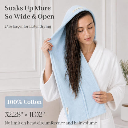 Woman in white robe using a light blue hair towel wrap draped over wet dark hair. Text states 'Soaks Up More, So Wide & Open,' '25% larger for faster drying,' '100% Cotton,' and dimensions 32.28" x 11.02".