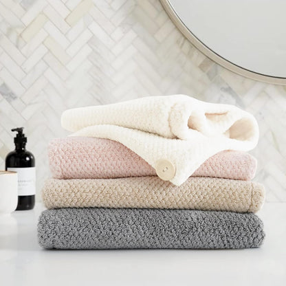 Stack of four fluffy textured towels in grey, beige, pink, and cream/white, folded and piled on a white surface in a bathroom setting with herringbone tile and a round mirror in the background.