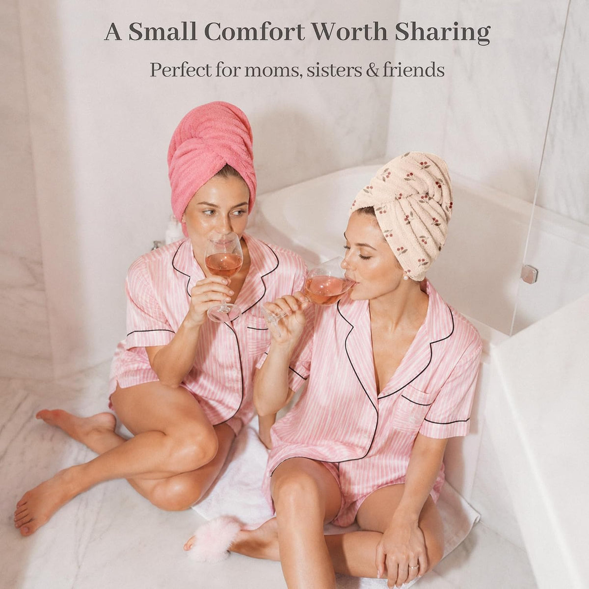 Two women in matching pink striped pajamas sit in a bathtub sipping rose wine, each wearing a hair towel turban -- one solid pink, one cream with a floral pattern. Text reads 'A Small Comfort Worth Sharing. Perfect for moms, sisters & friends.'
