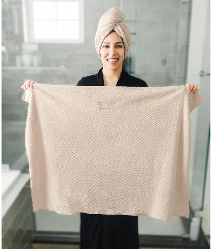 A smiling woman in a dark robe holds up a large light beige textured hair towel in a bathroom setting. She wears a matching hair turban wrap on her head, demonstrating both uses of the product.