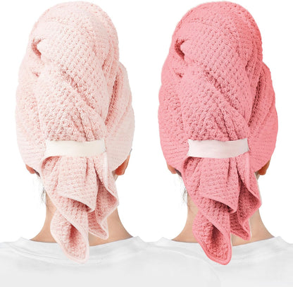 Two hair towel wraps worn on the backs of heads: one light pink and one coral pink. Both feature a waffle-knit texture, a white elastic band closure, and a draped tail section hanging down.