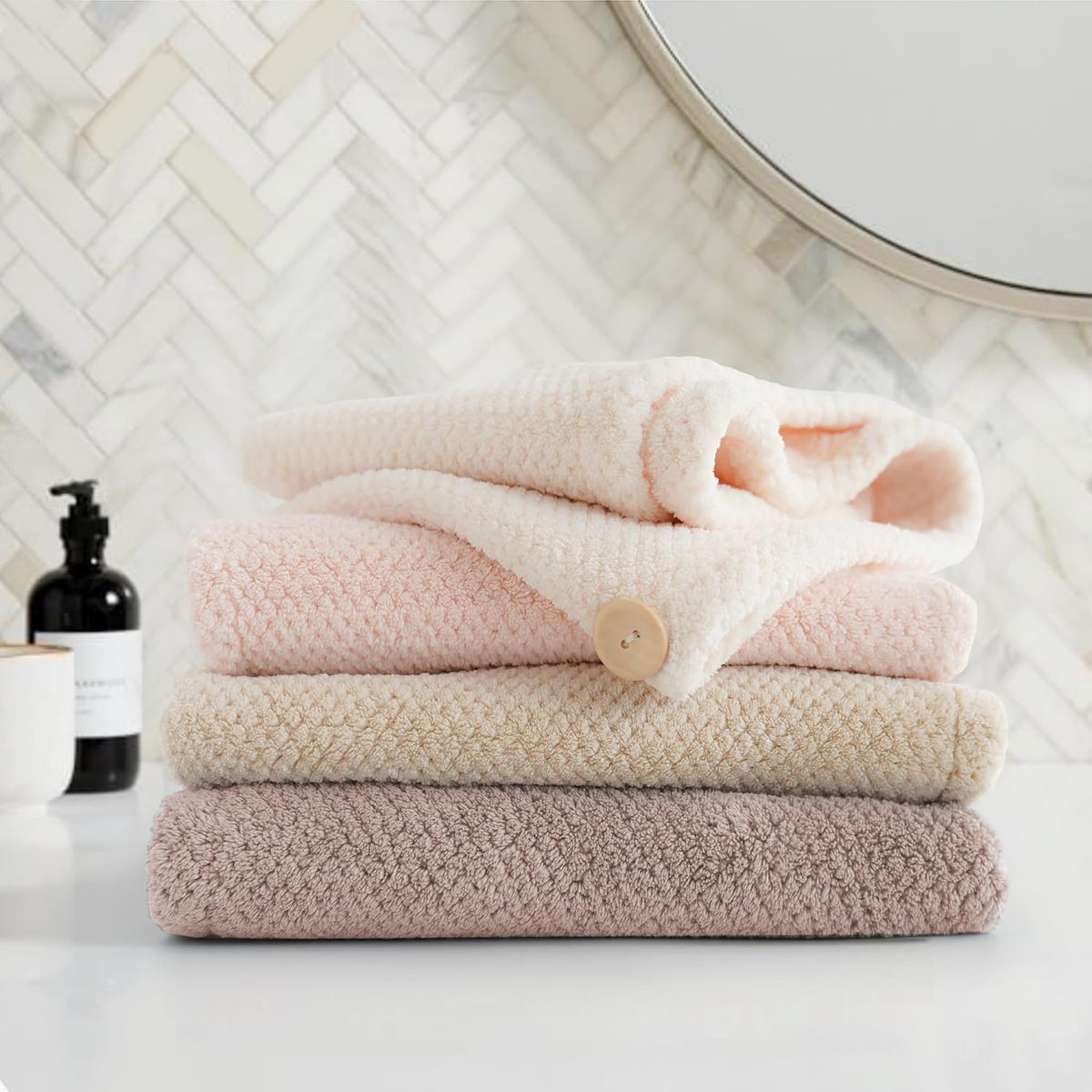 Stack of four folded textured towels in a bathroom setting: light pink on top, medium pink, cream, and dusty mauve at the bottom. A tan button detail is visible on the pink towel. A black soap dispenser sits nearby.