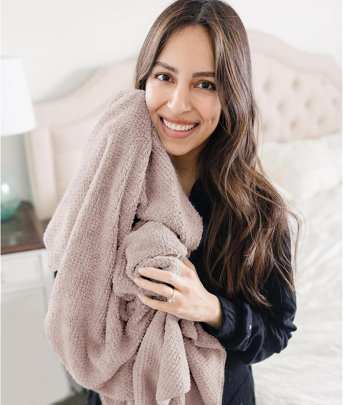 A smiling woman with long dark hair hugs a large, fluffy mauve-colored towel against her chest while wearing a dark top. She stands in a softly lit bedroom setting, cradling the plush, textured towel.
