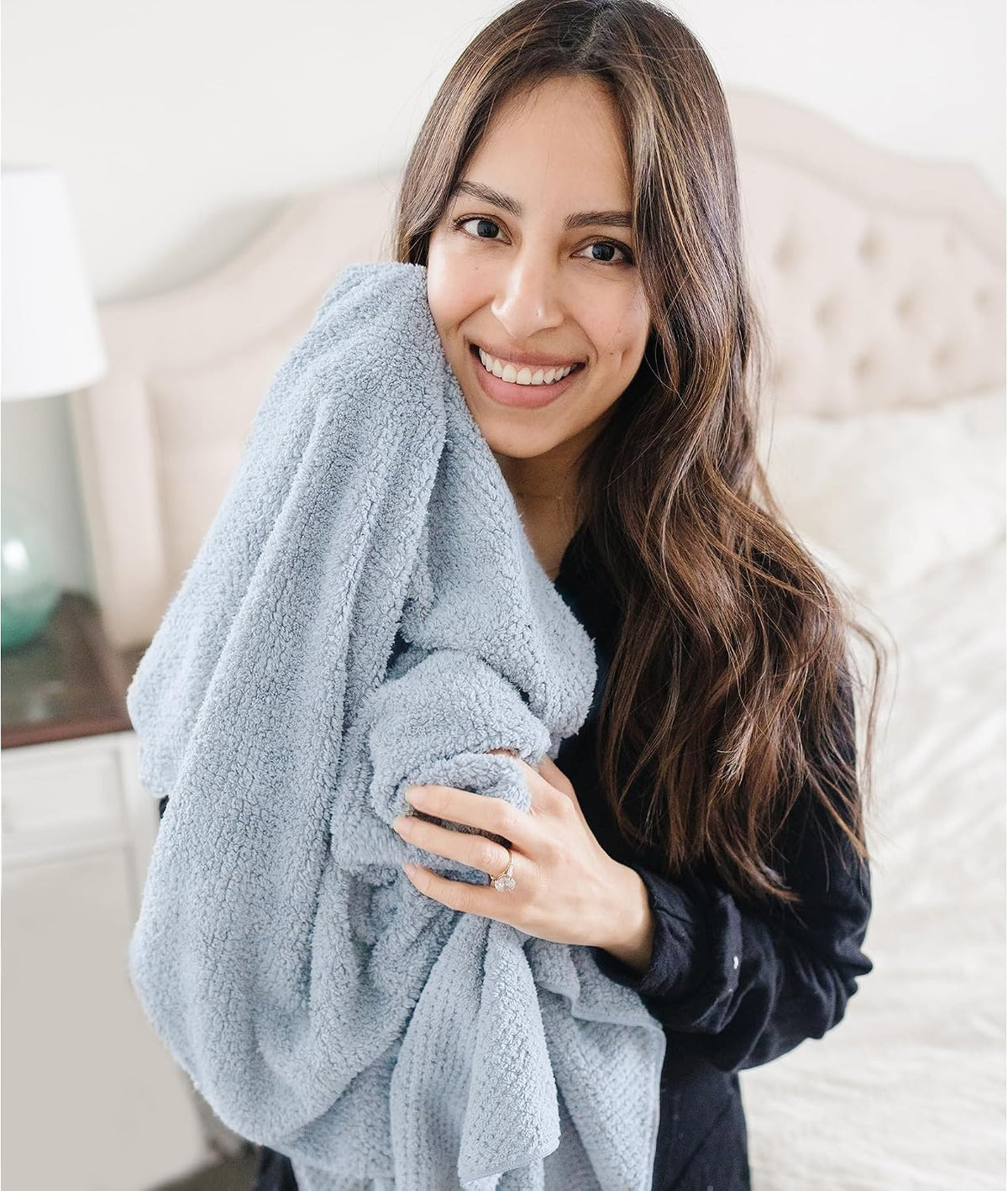 Woman with long dark hair smiling and hugging a large light blue fluffy textured towel against her chest, wearing a dark top. A bedroom with a tufted headboard is visible in the background.