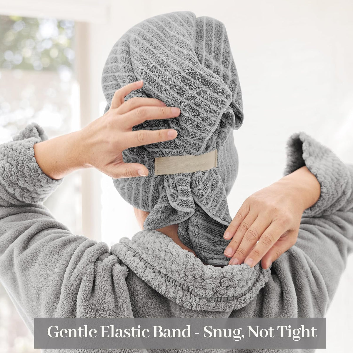 Person seen from behind wearing a grey textured hair towel wrap on their head, adjusting it with both hands. A beige elastic strap is visible at the back. Text overlay reads: Gentle Elastic Band - Snug, Not Tight.