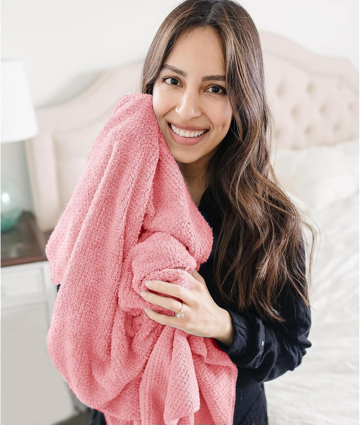 Woman in a dark top hugging a large coral pink textured towel against her cheek, smiling, in a bedroom setting. The soft, plush towel has a subtle waffle-like texture and is generously sized.