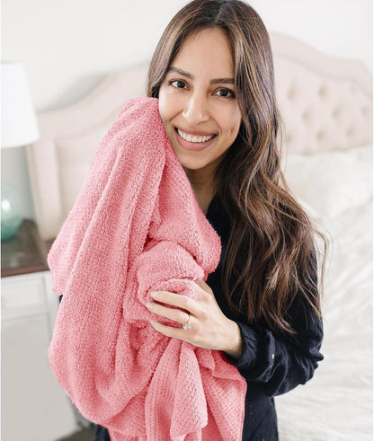 Woman in a dark top hugging a large coral pink textured towel against her cheek, smiling, in a bedroom setting. The soft, plush towel has a subtle waffle-like texture and is generously sized.