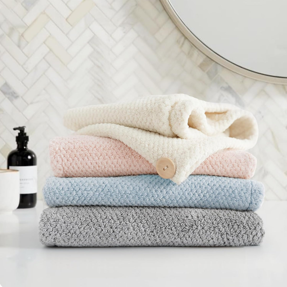 Stack of four folded textured towels in cream, pink, light blue, and gray, with a small wooden button visible on the pink towel. Arranged on a white surface in a bathroom setting with herringbone tile and a round mirror in the background.