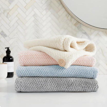 Stack of four folded textured towels in cream, pink, light blue, and gray, with a small wooden button visible on the pink towel. Arranged on a white surface in a bathroom setting with herringbone tile and a round mirror in the background.